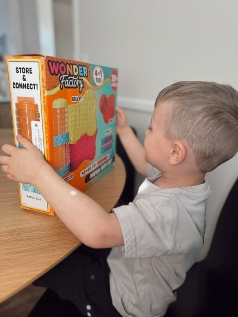 Toddler holding and exploring the Wonder Factory Never Dry Dough Mega Box Set on a wooden table, with the 4 in 1 tool packaging visible and colorful dough textures displayed on the box, available at Walmart.