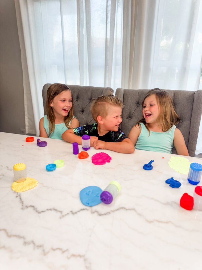 Alt text:
Three young children sitting at a table laughing and playing with colorful Wonder Factory Never Dry Dough, using rolling and stamping tools with bright dough pieces spread across the surface.