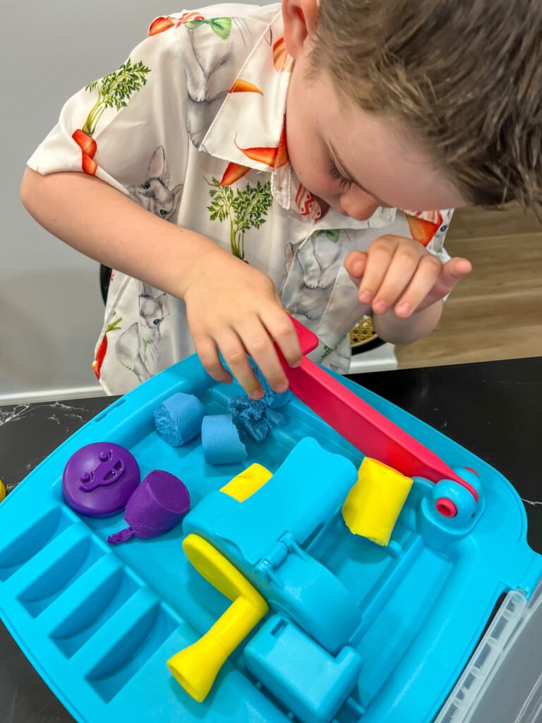 Close-up of a young child using the built-in rolling tool from the Wonder Factory Never Dry Dough set, shaping colorful gluten-free dough pieces inside the blue 4 in 1 activity tray.