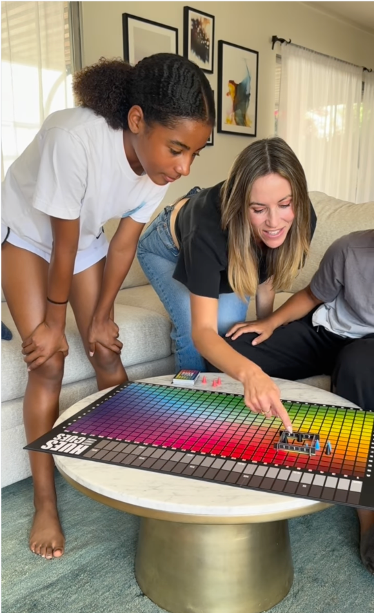Mom and two kids leaning over a coffee table playing Hues & Cues board game, pointing at the colorful grid board during family game night in a cozy living room.