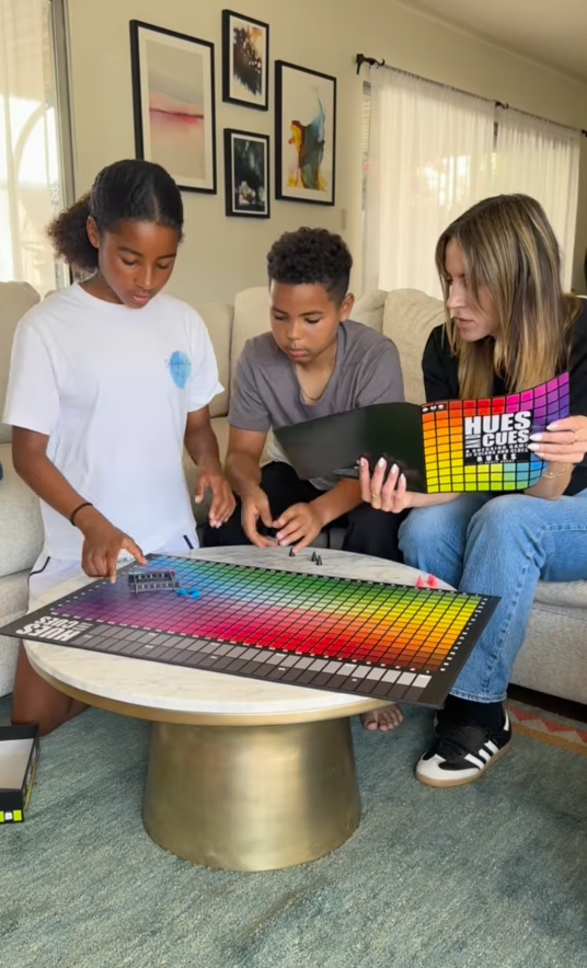 Mom and two children sitting on a couch playing Hues & Cues board game, placing pieces on the colorful grid board during a cozy family game night at home.