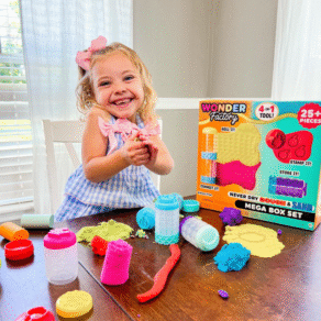 Smiling young girl playing with Wonder Factory Never Dry Dough Mega Box Set at a table, using colorful rolling and stamping tools, with gluten-free dough pieces spread out and product box visible in the background.
