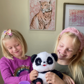 Two young girls smiling while playing with the My Baby Panda interactive toy from Little Live Pets, sitting at a table with the panda and its purple potty accessory, showcasing a fun Easter gift idea and screen-free indoor toy for kids.
