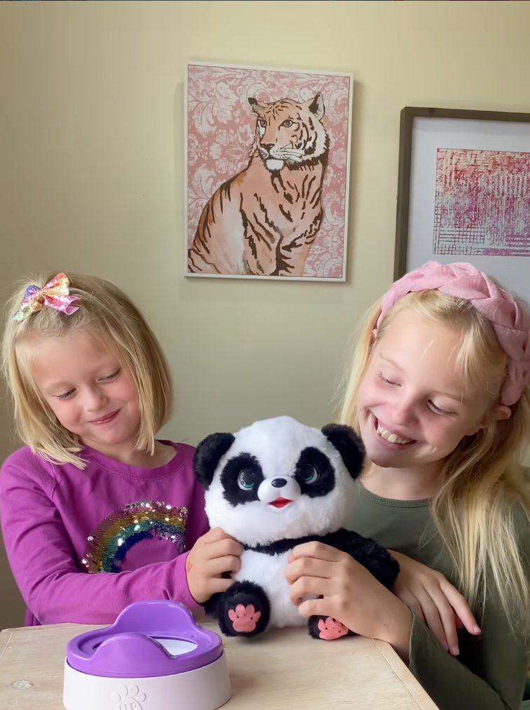 Two young girls smiling while playing with the My Baby Panda interactive toy from Little Live Pets, sitting at a table with the panda and its purple potty accessory, showcasing a fun Easter gift idea and screen-free indoor toy for kids.