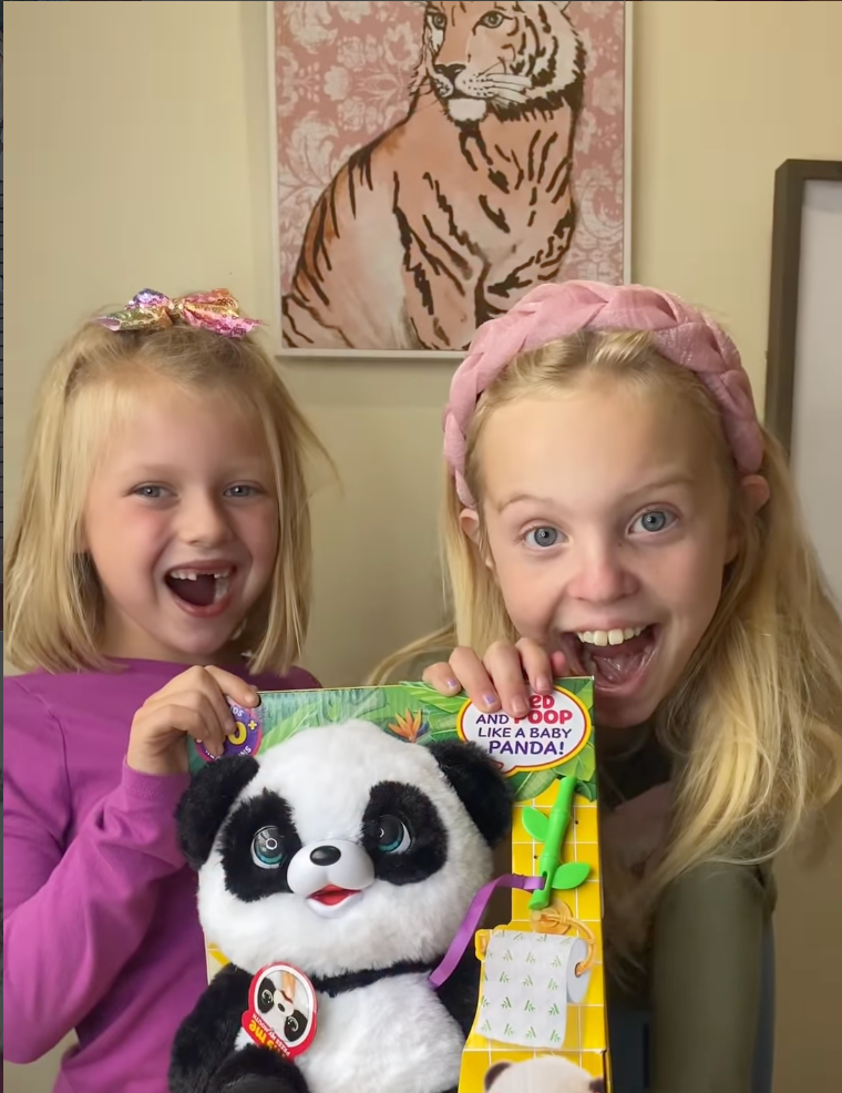 Two excited young girls holding the My Baby Panda interactive toy from Little Live Pets in its packaging, smiling and reacting with surprise, showcasing a fun Easter basket gift idea and interactive toy for kids.