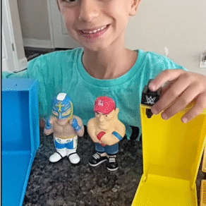 Child smiling while holding WWE Smash N Squish collectible figures next to colorful packaging cases on a kitchen counter
